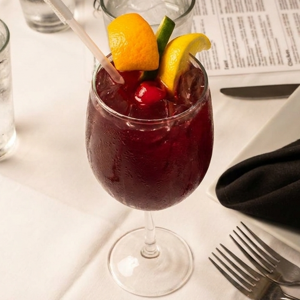 A red cocktail with orange, lemon, and cherry garnishes sits on a white tablecloth next to a water glass and menu.