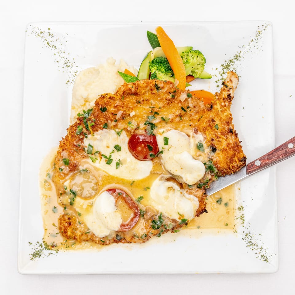 Breaded chicken cutlet topped with melted cheese and tomatoes, served with mashed potatoes and steamed vegetables on a white plate.