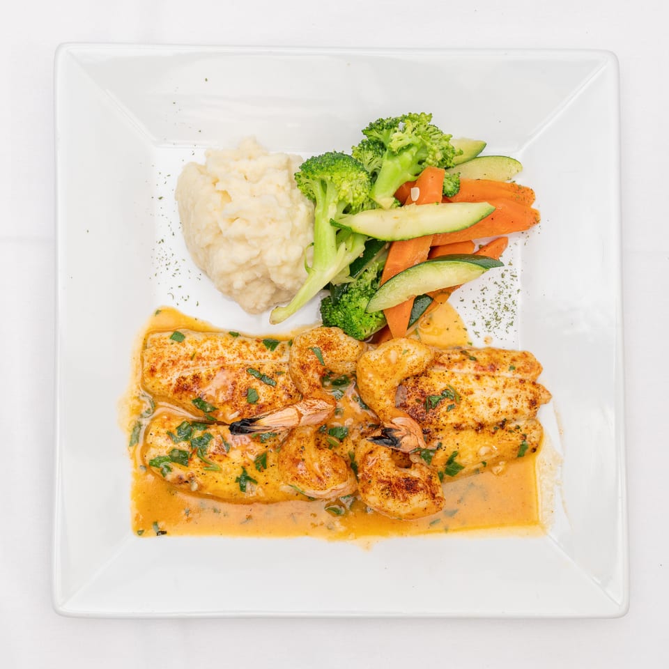 Creamy grilled chicken and shrimp with mashed potatoes and steamed broccoli, carrots, and zucchini on a white plate.