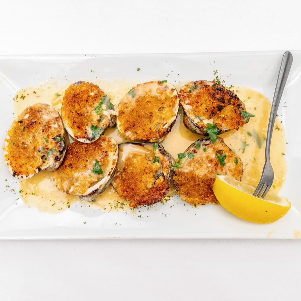 Baked stuffed clams topped with golden breadcrumbs and parsley, served in lemon butter sauce with a lemon wedge.