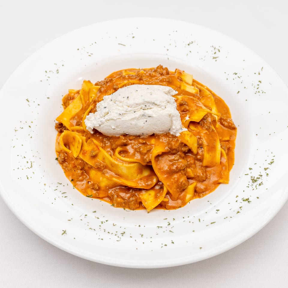Tagliatelle pasta in creamy meat sauce topped with a mound of seasoned ricotta on a white plate.