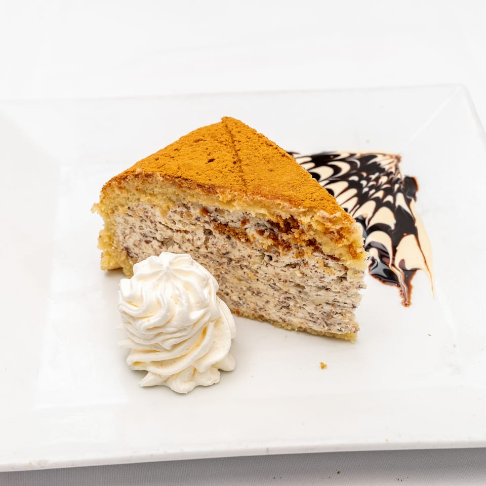 Slice of layered cake dusted with cinnamon, served with whipped cream and chocolate sauce on a white plate.
